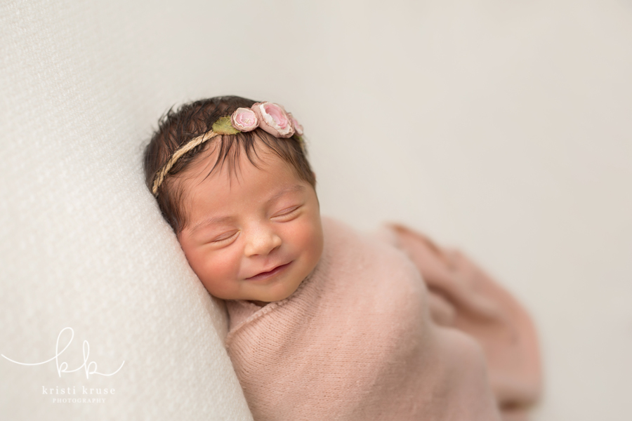 Newborn baby girl wrapped in pink blanket laying on white blanket with big smile on her face