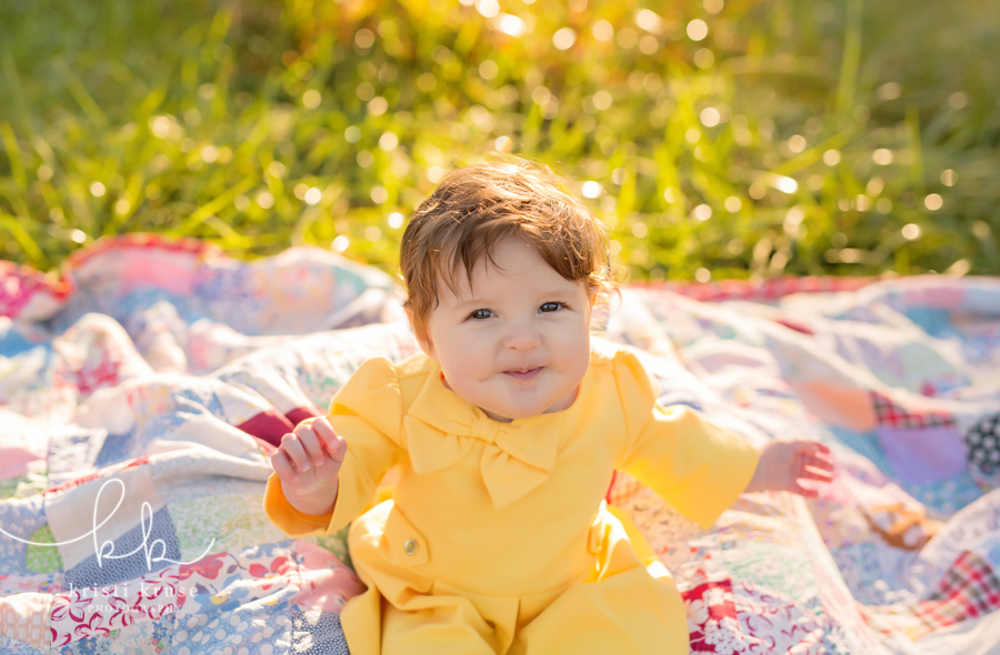 6 month old baby girl in yellow dress sitting on patchwork quilt in tall sunlit grass
