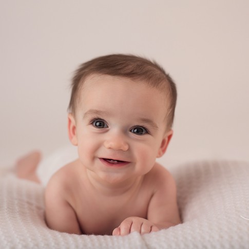 6 month old baby boy laying on belly on white blanket looking at camera