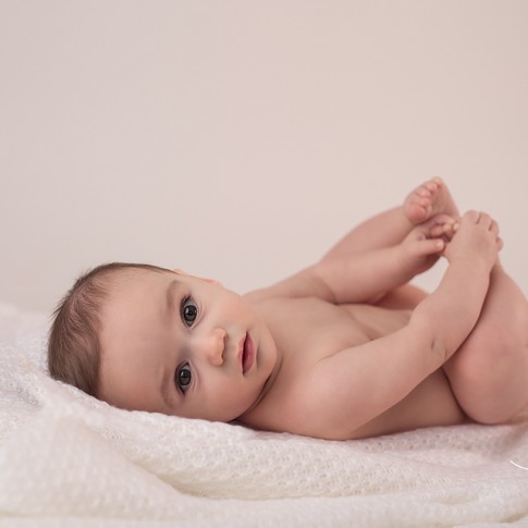 6 month baby lying on side on white blanket holding his feet looking at camera