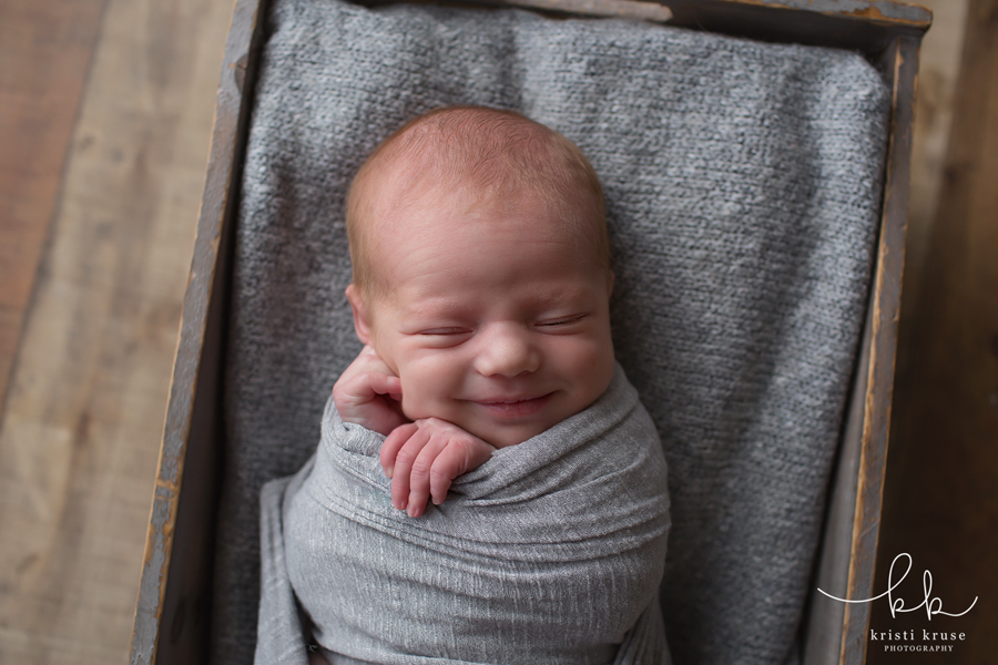 newborn baby boy wrapped in gray and laying in a gray box with gray blanket with big smile