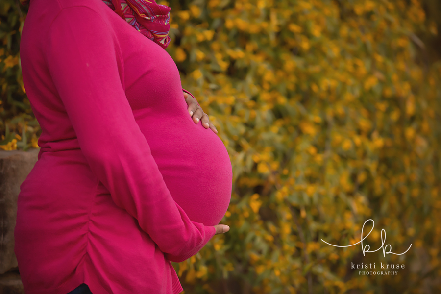 Pregnant woman standing sideways wearing bright pink sweater holding her belly in front of yellow flowering bush