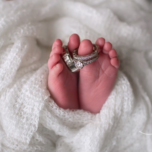 Newborn feet wrapped in white blanket with parents weddings rings on the toes