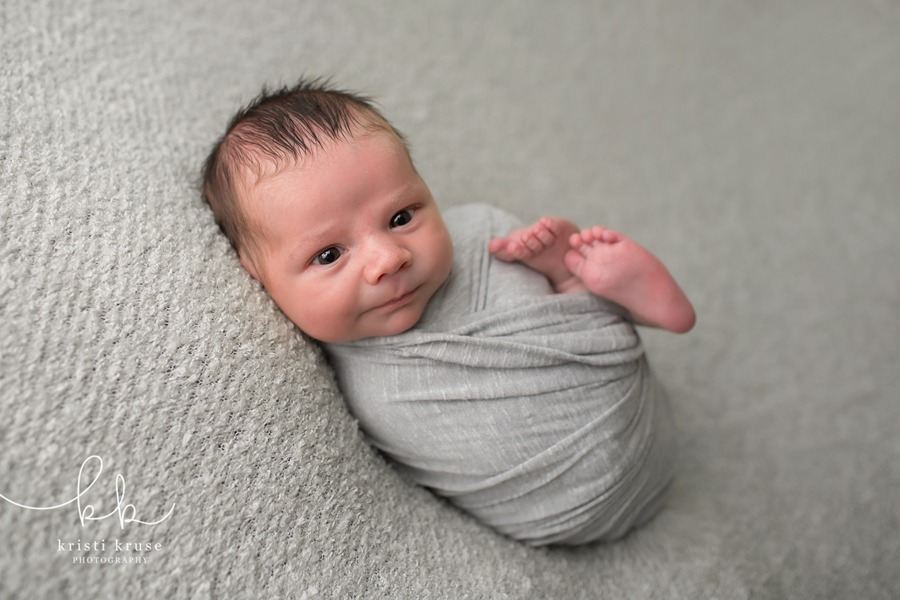 Newborn baby boy wrapped in gray swaddle laying on gray blanket smiling at the camera