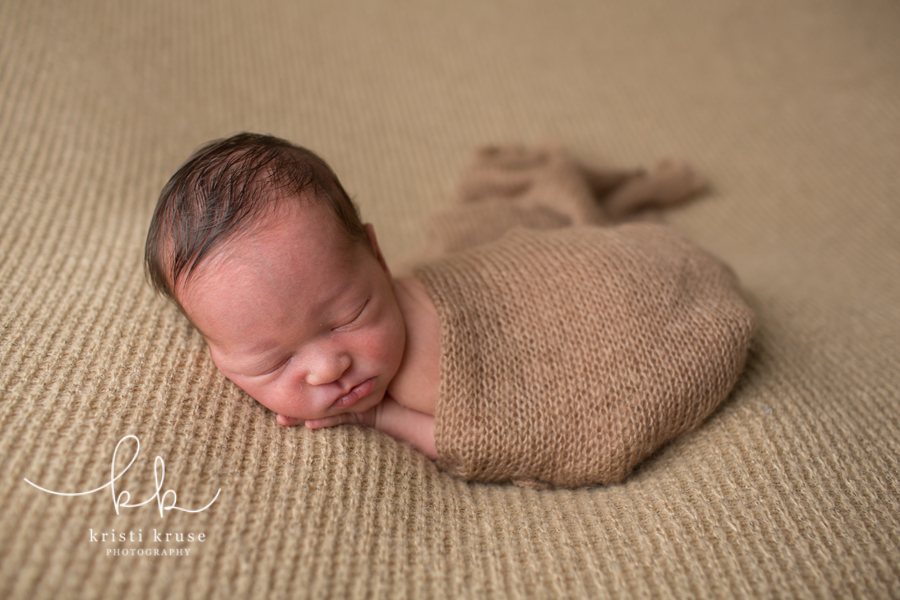 Newborn baby boy swaddled in brown wrap laying on his tummy on a brown blanket