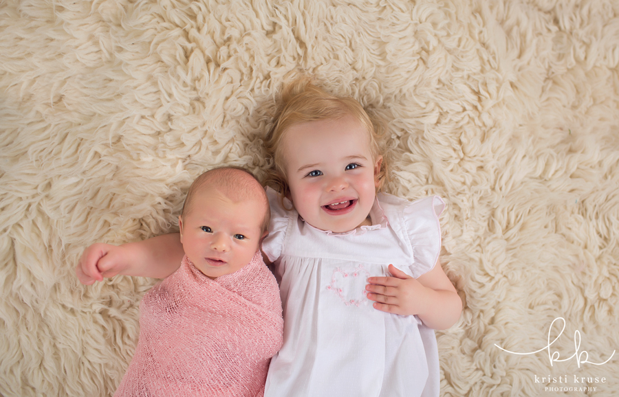 2 year old blond girl wearing white dress laying on cream rug while baby sister wrapped in pink swaddle lays next to her looking at camera