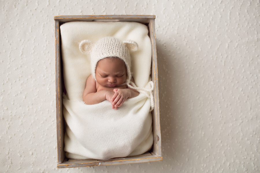 newborn baby girl in small bed with cream blanket and cream knit teddy bear hat