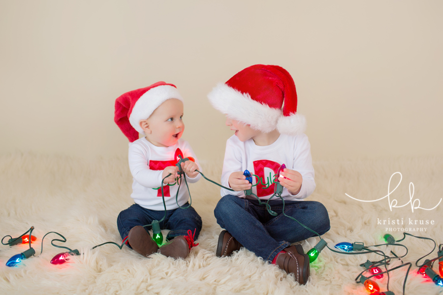 3 year old and 6 month old brothers wearing white shirts with their initials and blue jeans with santa hats and christmas lights