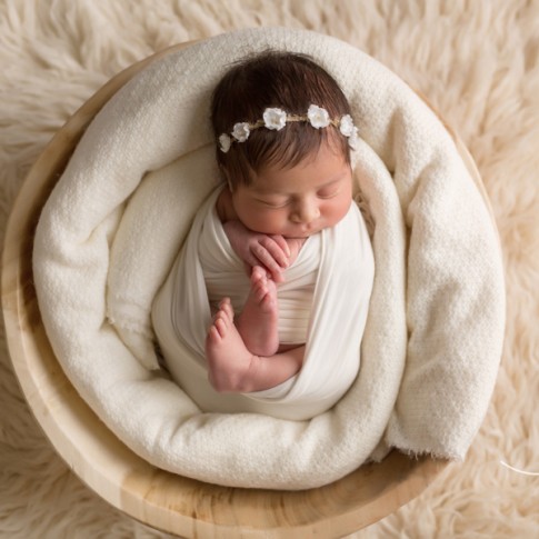 newborn baby girl wrapped in white swaddle laying in wooden bowl on