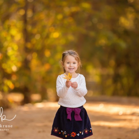 3 year old girl with ponytail wearing white shirt and blue skirt holding a leaf on a wooden trail