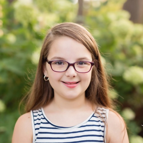 9 year old girl with long brown hair and glasses in front of hydrangea bush