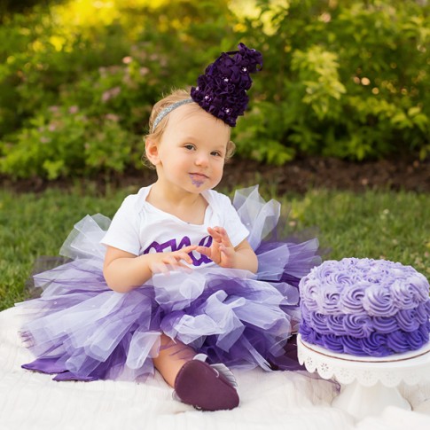 1 year old girl wearing purple tutu with matching hat and onesie with purple birthday cake