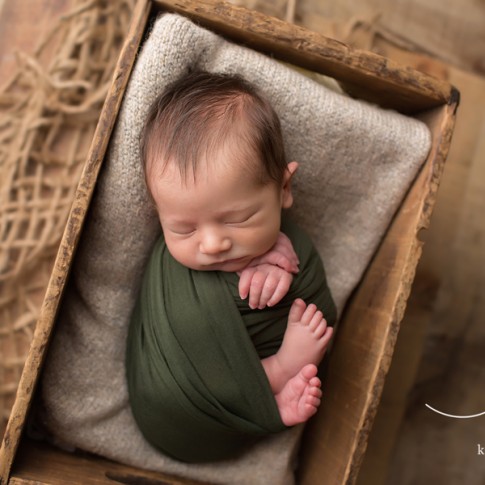 baby boy wrapped in olive swaddle laying in wooden crate with gray blanket