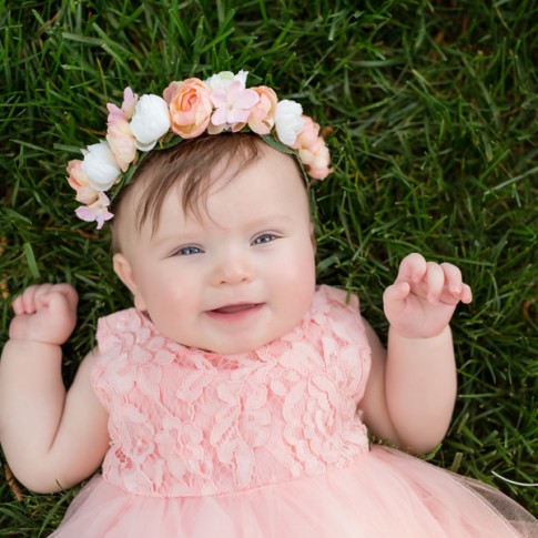 6 month old girl wearing pink dress with pink flower crown laying on grass