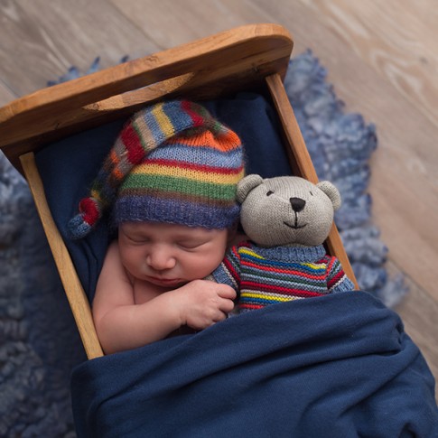 Baby boy with striped hat laying in smal wooden bed with teddy bear in matching sweater