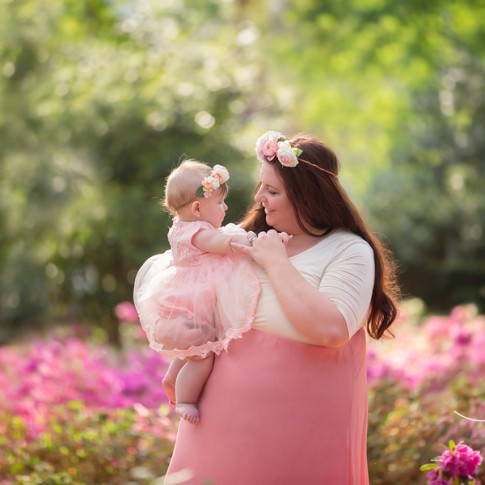 Mother holding 6 month old daughter wearing flower crown. Both are standing in a flower garden